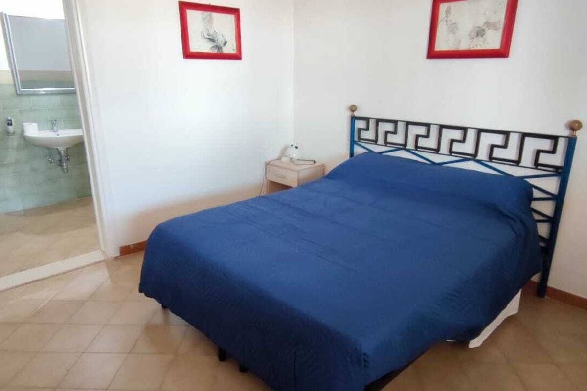 Quarto com cama, mesa de cabeceira e casa de banho visível através de uma porta de vidro.