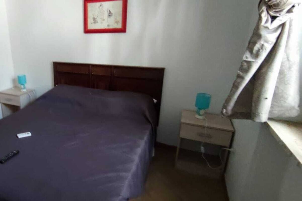 Uma cama de solteiro com colcha escura, cabeceira de madeira, mesa de cabeceira com abajur e um quadro na parede.