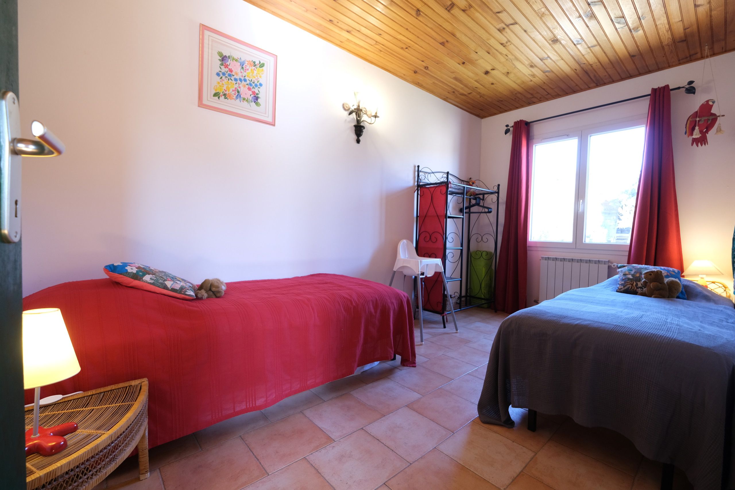 Une chambre avec deux lits, une chaise en osier, un radiateur, une fenêtre avec des rideaux et un luminaire fixé au mur.
