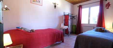 A room with two beds, a wicker chair, a radiator, a window with curtains, and a wall-mounted light fixture.