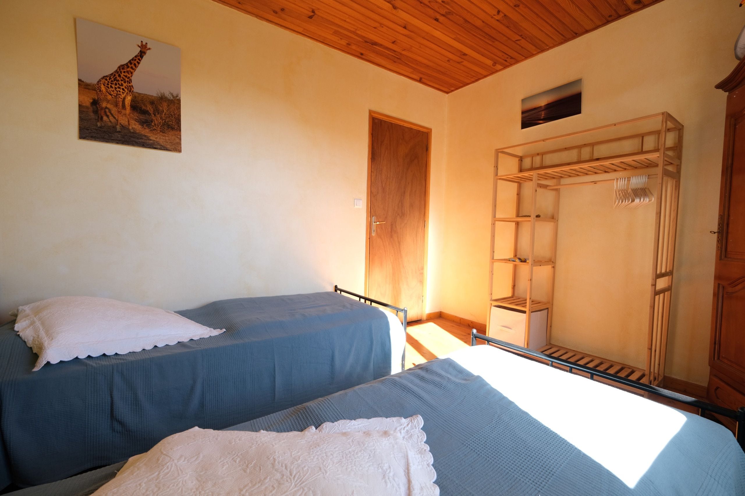 Ein Schlafzimmer mit zwei Einzelbetten, einer Holzbalkendecke, einer Tür, einem Regal und einem Giraffenbild an der Wand.