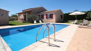 Una piscina con escaleras metálicas, un patio con sillas de descanso y una casa con contraventanas rojas.