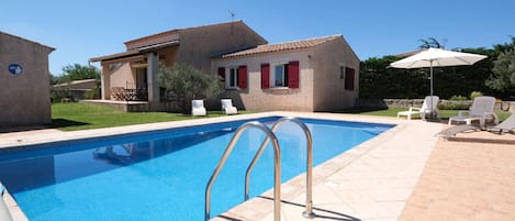 A swimming pool with metal ladders, a patio with lounge chairs, and a house with red shutters.