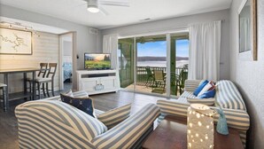 Ein geräumiges Wohnzimmer mit großem Fernseher, gestreifter Couch, Essbereich und Balkon mit Seeblick.