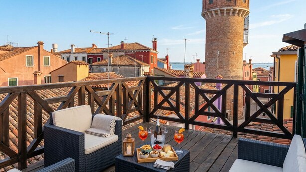 Una terrazza con mobili in vimini, un tavolo con snack e bevande, e vista su edifici colorati e una torre.