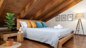 Una camera da letto mansardata accogliente, con soffitto in legno, un letto con cuscini bianchi e colorati, una pianta sul comodino, una lampada da terra e stampe botaniche incorniciate alle pareti.