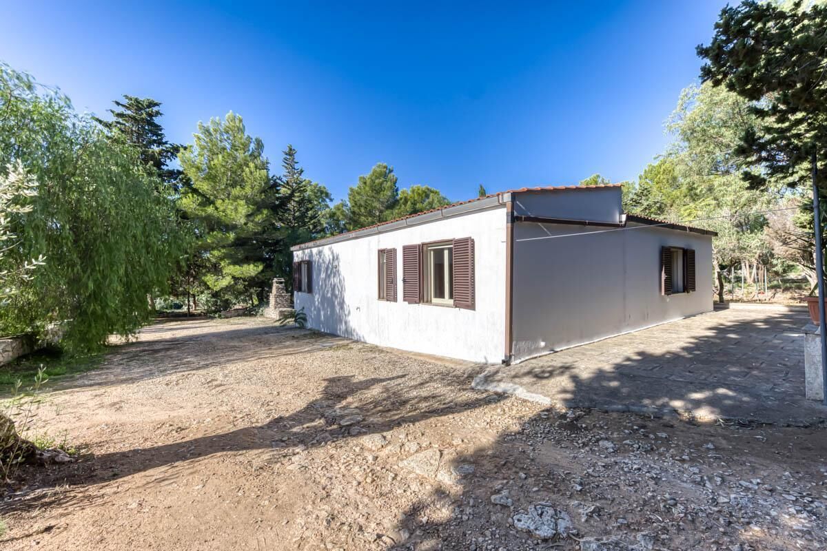 A single-story building with a white exterior and brown shutters, surrounded by trees and a gravel driveway.