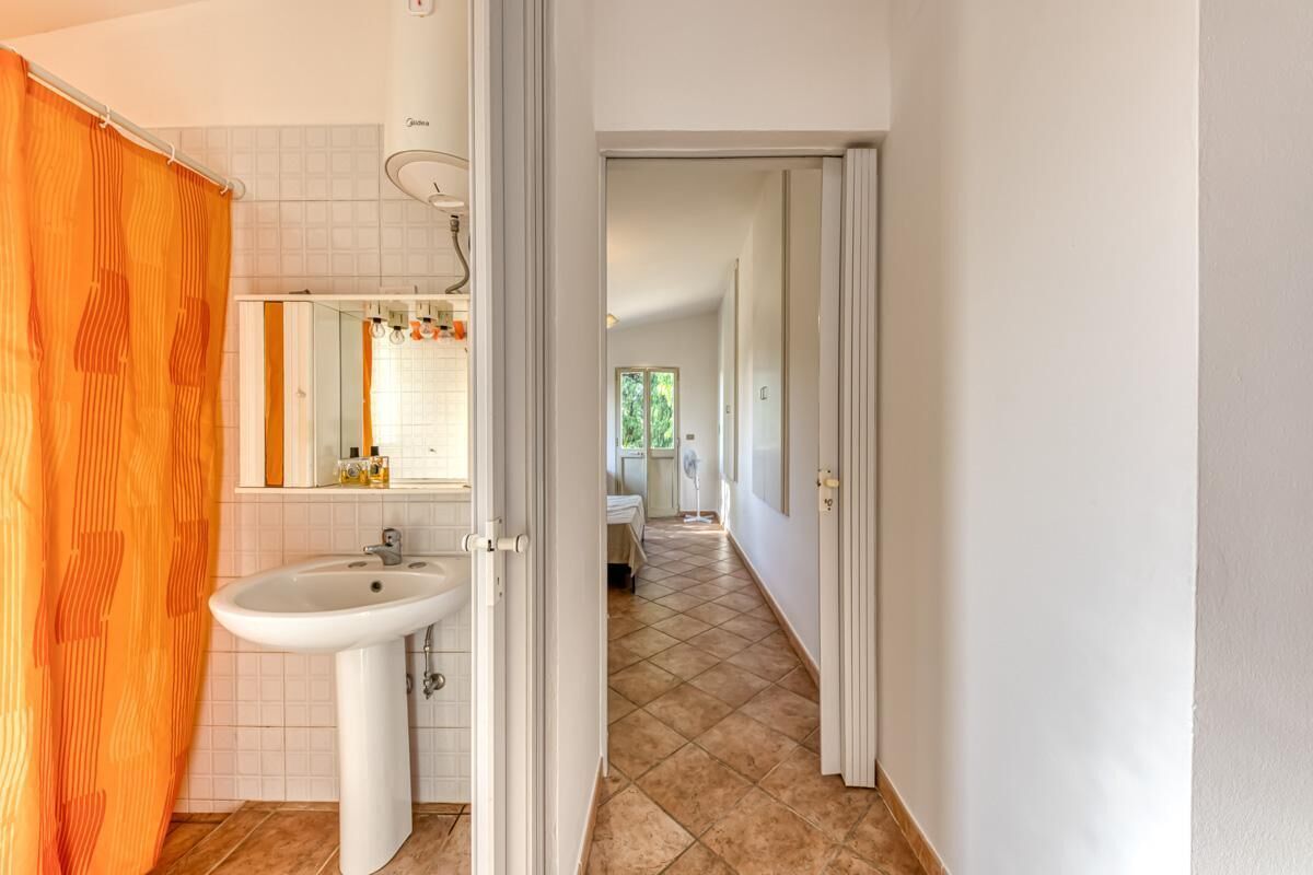 A bathroom with a sink, mirror, and orange shower curtain. A hallway leads to a bedroom with a bed and window.