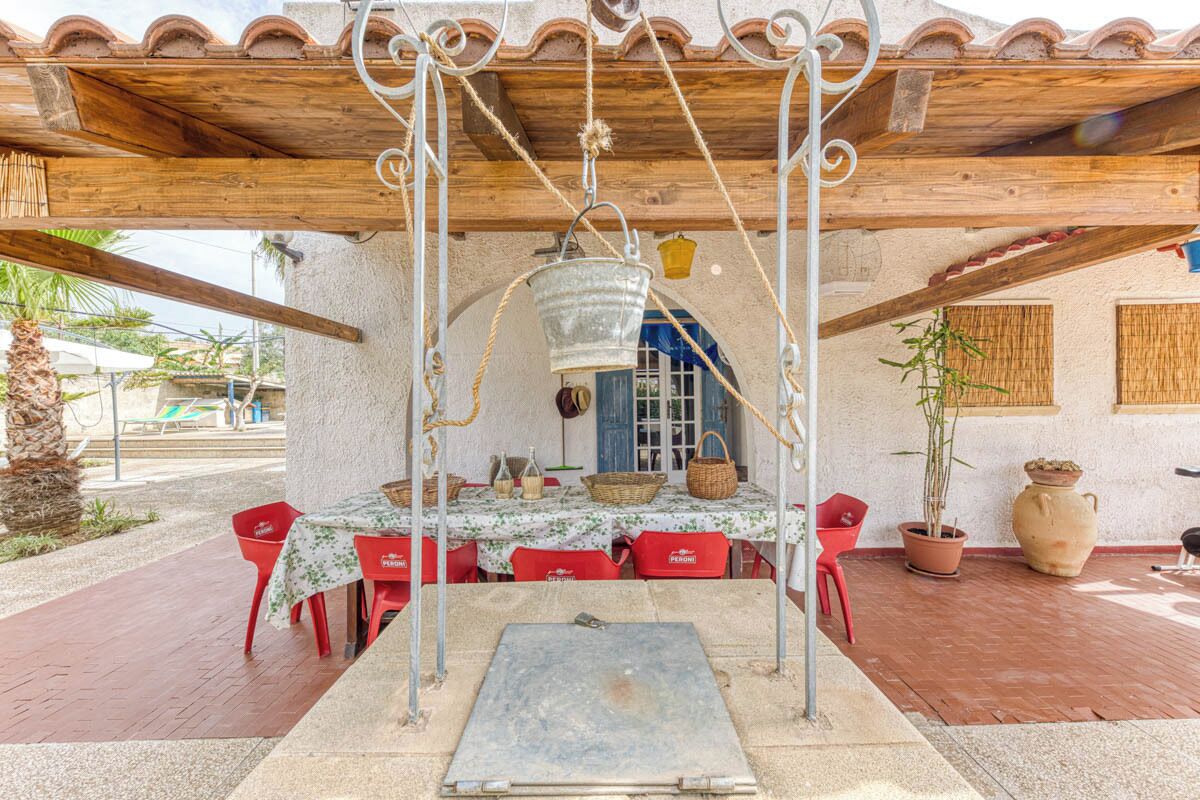 Un patio al aire libre con una pérgola de madera, una mesa con mantel floral, sillas rojas y plantas en macetas.