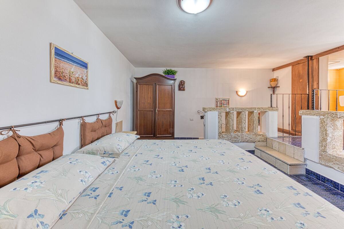Een slaapkamer met een groot bed, een houten garderobe, een klein tafeltje met een plant en een badkamer met een betegelde vloer en een douchehoek.