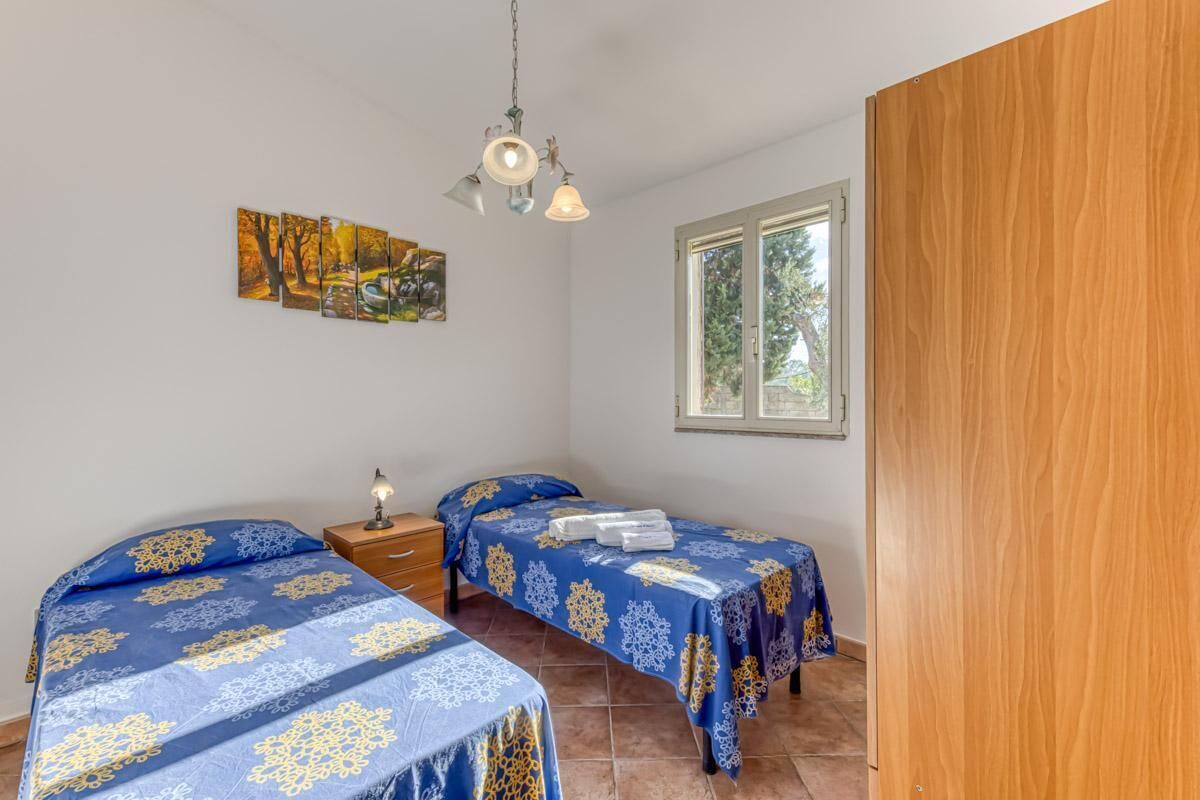A bedroom with two beds, a nightstand, a hanging light fixture, and a window with a view of trees.