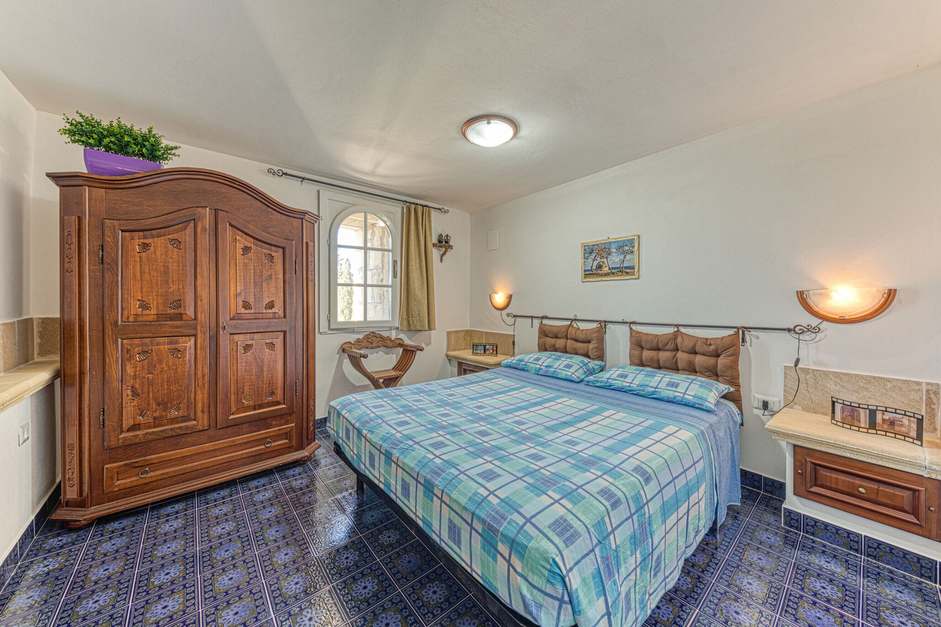 Un dormitorio con un armario de madera grande, una cama con colcha azul de cuadros, una ventana con cortina y una estantería con un marco de fotos.