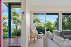 A modern interior with a sofa, a chair, and a large sliding glass door leading to a balcony with a view of palm trees and the ocean.