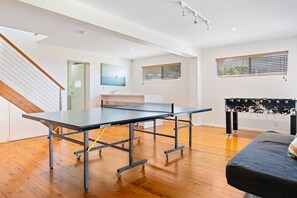 Una habitación con mesa de ping pong, suelo de madera, una cama, un ventanal con persianas y un cuadro en la pared.