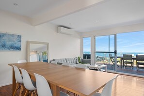 Um quarto espaçoso com mesa de jantar de madeira, cadeiras brancas, sofá cinzento e uma grande janela com vista para o oceano.