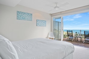 A bedroom with a large bed, a ceiling fan, and a view of the ocean through a sliding glass door.