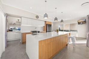 A modern kitchen with a central island, wooden cabinets, stainless steel appliances, pendant lighting, and a large window. - Sea Sand Serenity-Stunning Sophistication Stockton (Stockton)