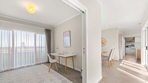Una sala de estar moderna y bien iluminada, con un ventanal grande, un comedor con mesa y sillas, y una cocina visible a través de un umbral abierto.