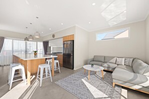 Een moderne keuken met een centraal eiland, een inbouwoven en een koelkast. Er is een zithoek met een witte tafel en twee witte krukken. Een grijze bank staat in de woonkamer met een vloerkleed met een patroon.