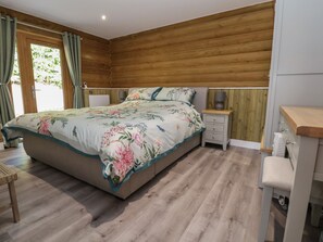 Un dormitorio con cabecera de madera, colcha estampada, mesita de noche con lámpara y una cómoda pequeña.