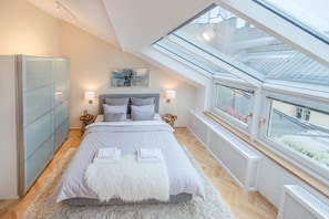 A bedroom with a skylight, a bed with pillows and a fluffy throw, a bedside table with a lamp, and a radiator.
