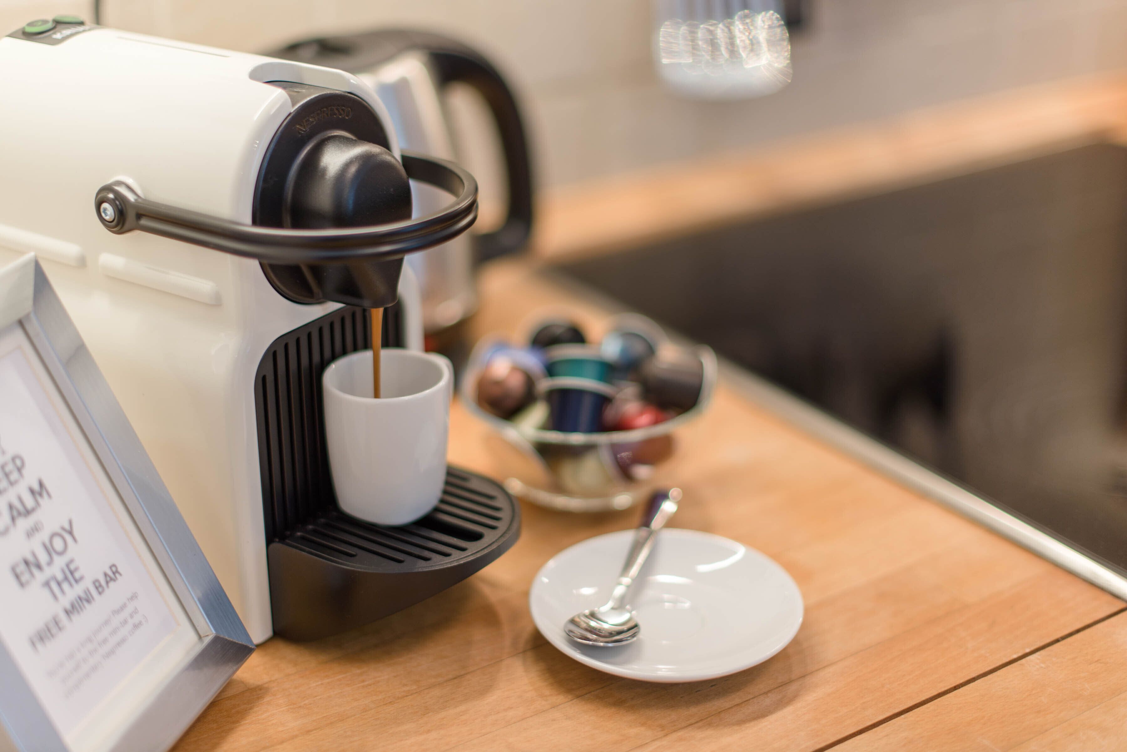 Una cafetera sirviendo café en una taza, un plato con una cuchara y un tazón de cápsulas de café sobre una mesa de madera.