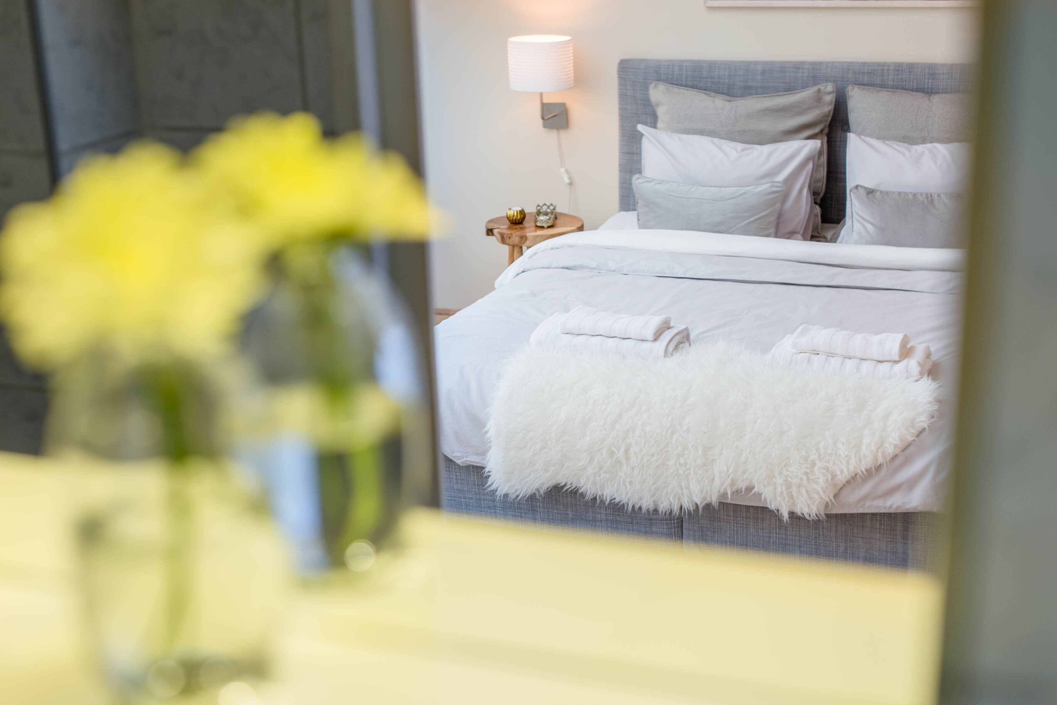 A neatly made bed with white linens and a fluffy white throw blanket, a bedside lamp, and a small wooden nightstand with a decorative item.