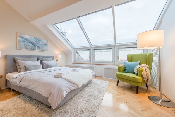A bedroom with a skylight, a bed with white bedding, a green armchair, and a floor lamp.