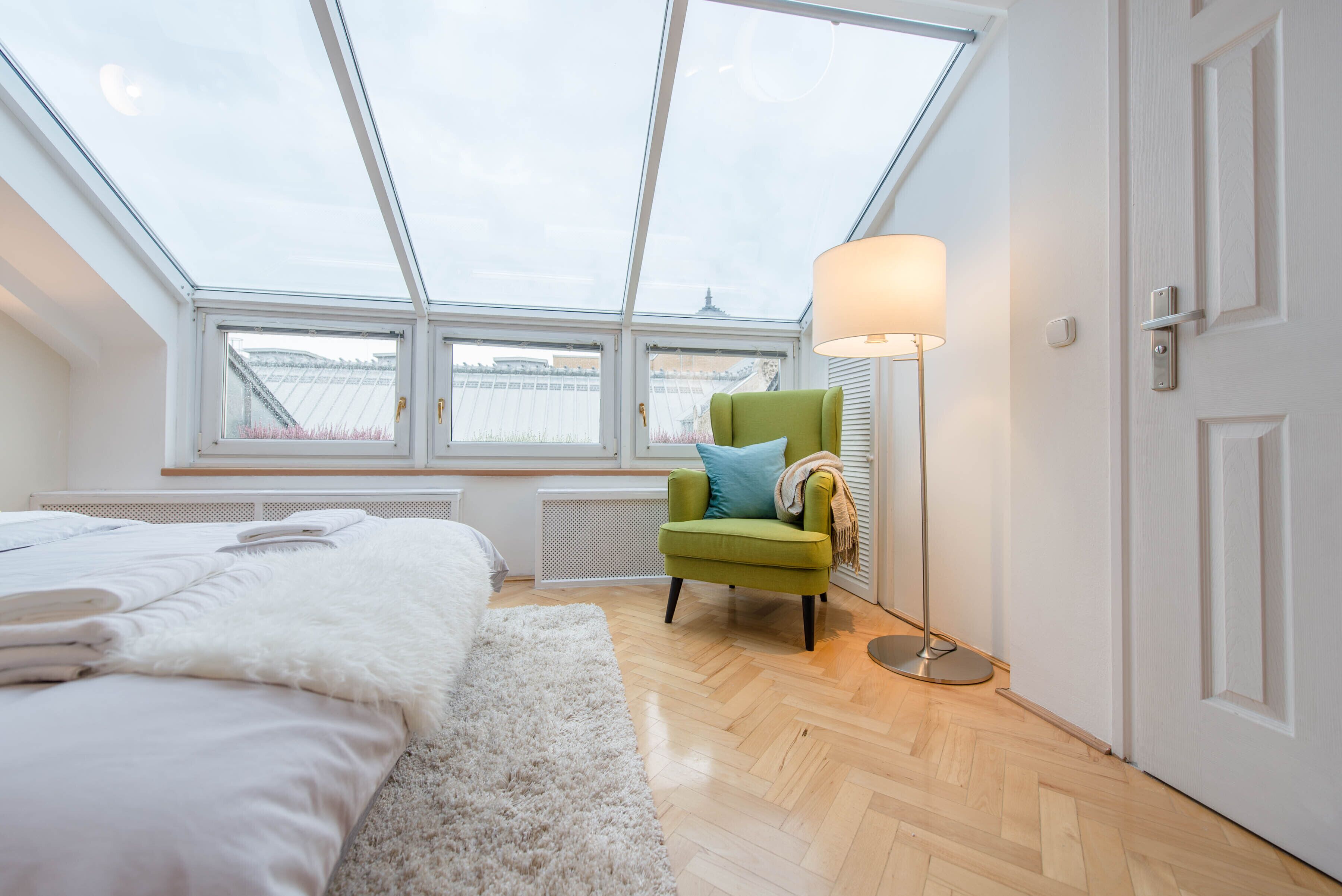 A bedroom with a skylight, a green chair, a floor lamp, and a bed with white bedding.
