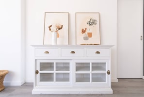 A white cabinet with three drawers and a glass-fronted section, displaying two framed artworks above it.