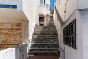 A narrow stairway with a metal railing, leading to a building with a white facade and a window with a barred design.