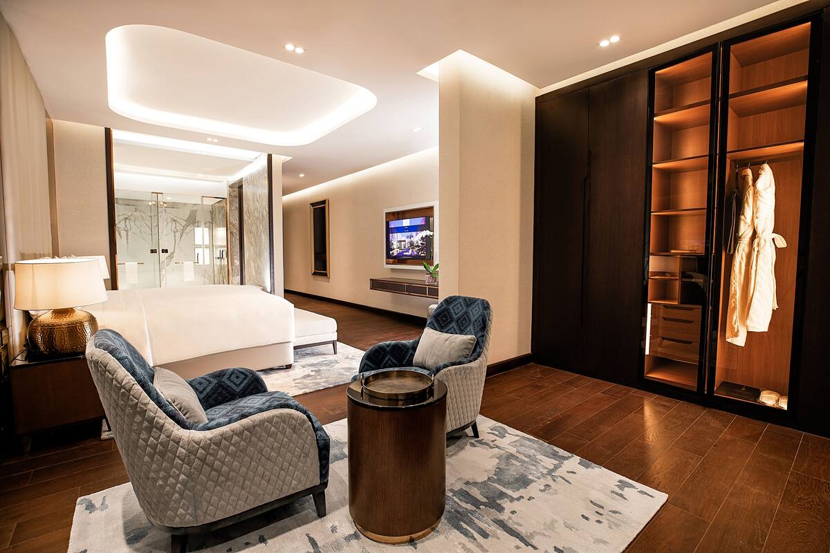 Ein modernes Hotelzimmer mit einem großen Bett, zwei Sesseln, einem Couchtisch, einem Fernseher und einem begehbaren Kleiderschrank.