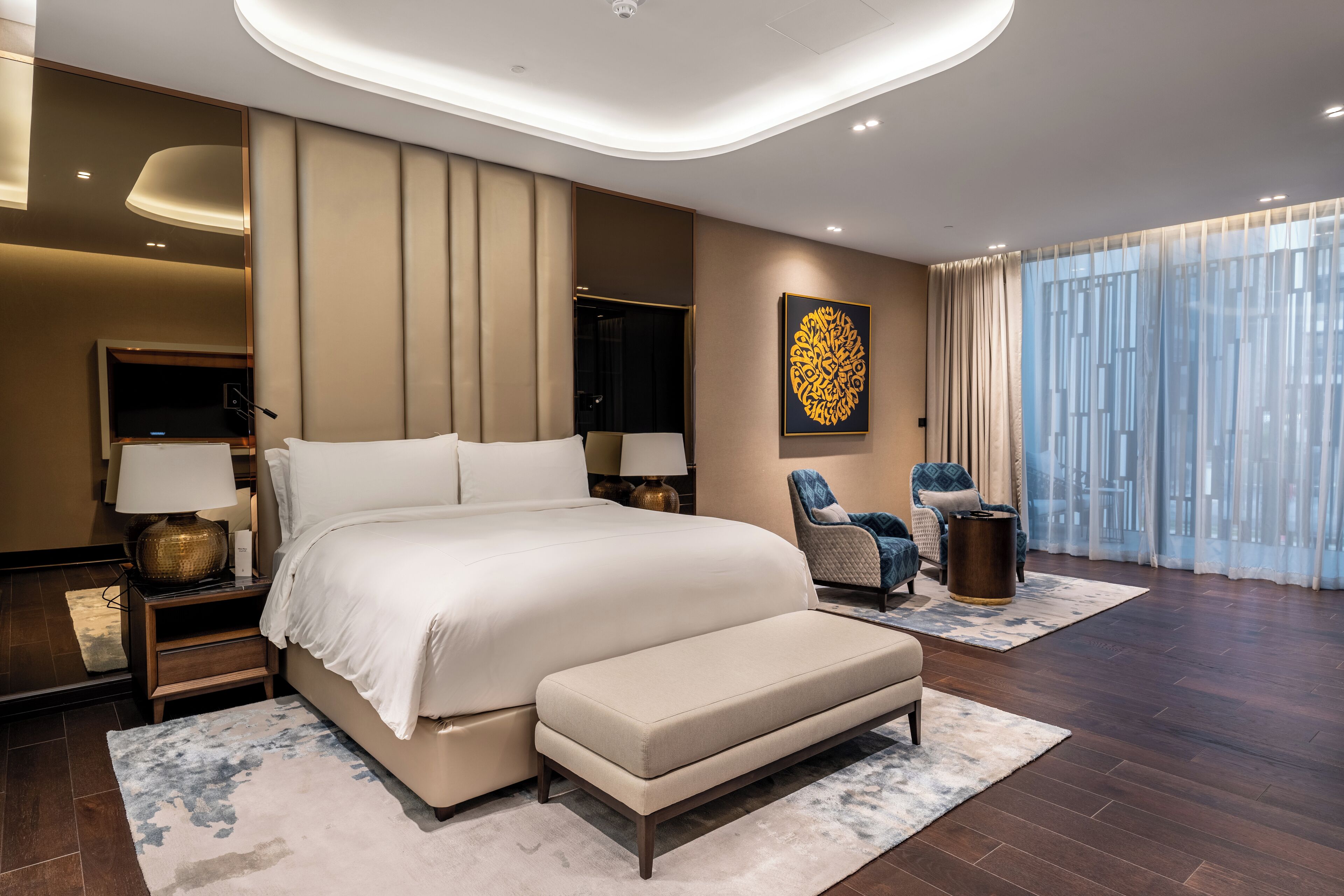 A modern bedroom with a large bed, bedside tables, a bench, a chair, and a large window with curtains.