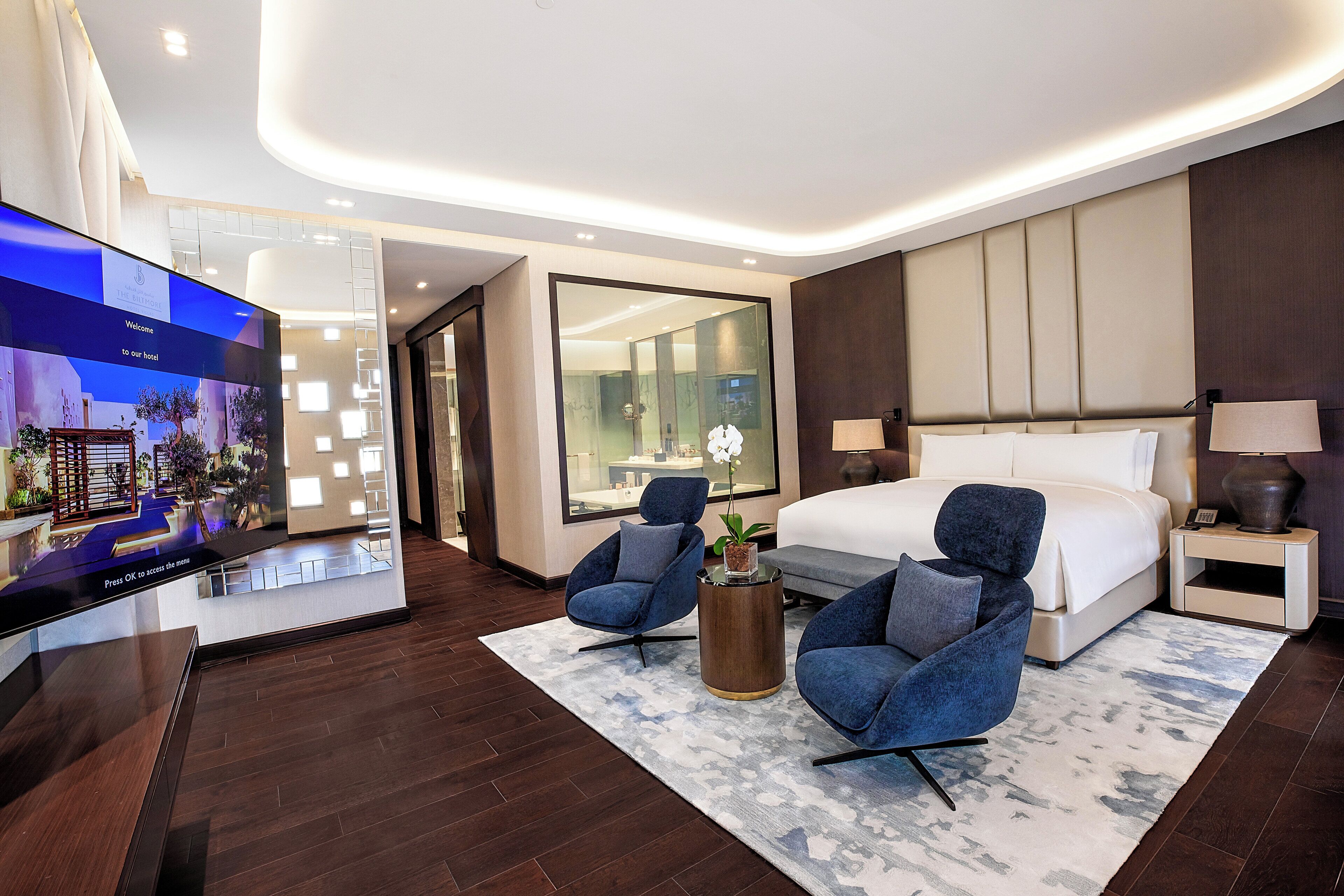 Ein modernes Hotelzimmer mit einem großen Bett, zwei Sesseln, einem Fernseher und einem großen Fenster.