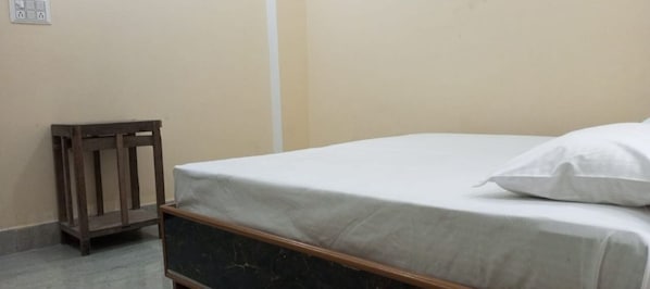 A single bed with a white mattress and pillow in a room with beige walls and a wooden headboard.