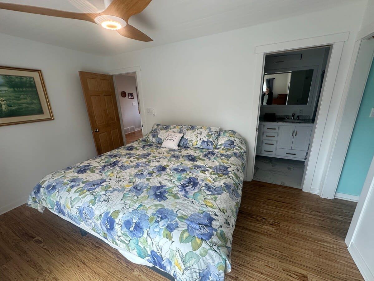 Une chambre avec un plancher en bois, un lit recouvert d’une literie à motifs floraux, un ventilateur de plafond et une porte donnant sur une salle de bain.