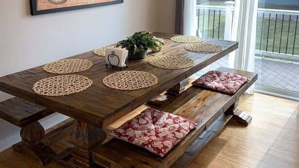 Una mesa de comedor con individuales tejidos, bancos con cojines estampados, un cuadro en la pared y vistas al exterior a través de un ventanal.
