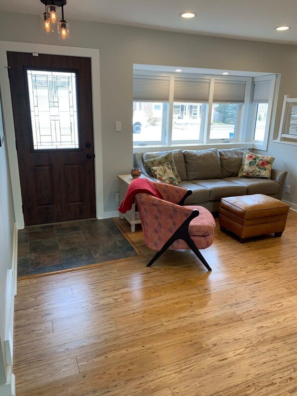 Un salon avec un plancher en bois, une porte marron, une fenêtre avec des stores, un canapé beige, un fauteuil à motifs et un pouf marron.