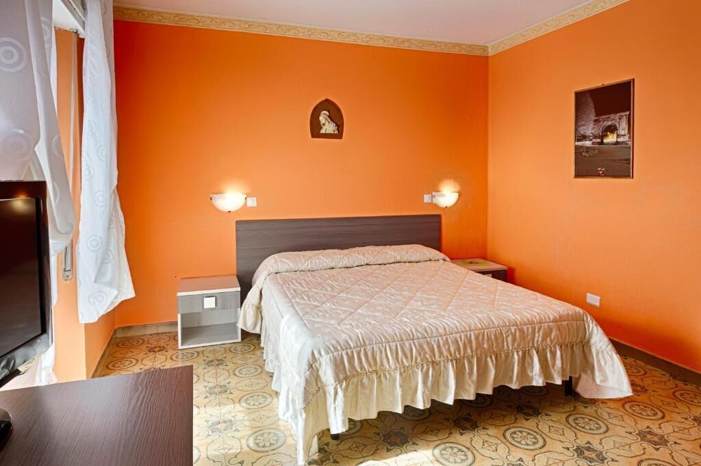 Una camera da letto con una parete arancione, un letto con copriletto beige, due lampade da comodino, un comodino piccolo, una TV e un pavimento a motivi.