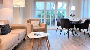 Ein modernes Wohnzimmer mit Sofa, Sessel und Couchtisch. Es gibt einen Essbereich mit Tisch und Stühlen. Große Fenster sorgen für natürliches Licht und einen Blick ins Freie.