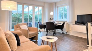 Ein modernes Wohnzimmer mit Sofa, Esstisch mit Stühlen, Fernseher und großen Fenstern mit Vorhängen.