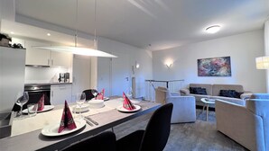 Eine moderne Küche mit weißen Schränken, eine Essecke mit einem langen Tisch für vier Personen und ein Wohnbereich mit einer Couch und einem Bild an der Wand.