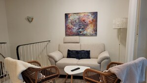 Ein gemütliches Wohnzimmer mit einer Couch, zwei Korbstühlen, einem kleinen weißen Tisch und einem farbenfrohen abstrakten Gemälde an der Wand.