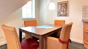 Een eetgedeelte met een houten tafel en vier rieten stoelen, een schilderij van Mexicaanse tortilla's en een raam met natuurlijk licht.