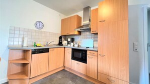 Een kleine keuken met houten kastjes, een magnetron, een koffiezetapparaat en een klok aan de muur.