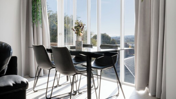Un comedor moderno con una mesa y sillas, ventanas amplias con cortinas y vistas a la ciudad.
