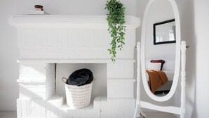 Una chimenea de ladrillo blanco con una planta colgante, una cesta de mimbre blanca y un espejo blanco con marco negro.