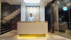 A reception desk with a person standing behind it, a staircase to the left, and a glass door to the right. - Hotel Madhavam (Mathura)