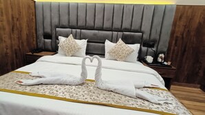 A neatly made bed with two swan-shaped towels, a floral-patterned bedspread, and a headboard with a vertical panel design. - Hotel Madhavam (Mathura)