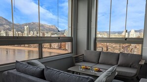 Une chambre moderne avec vue sur la ville, un canapé, une petite table avec des boissons et une fenêtre donnant sur un plan d’eau et des montagnes.
